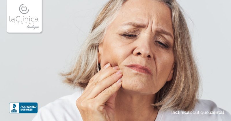 Mujer mayor con dolor dental, tocándose la mejilla, representando la preocupación por la salud dental en La Clínica Dental Boutique.