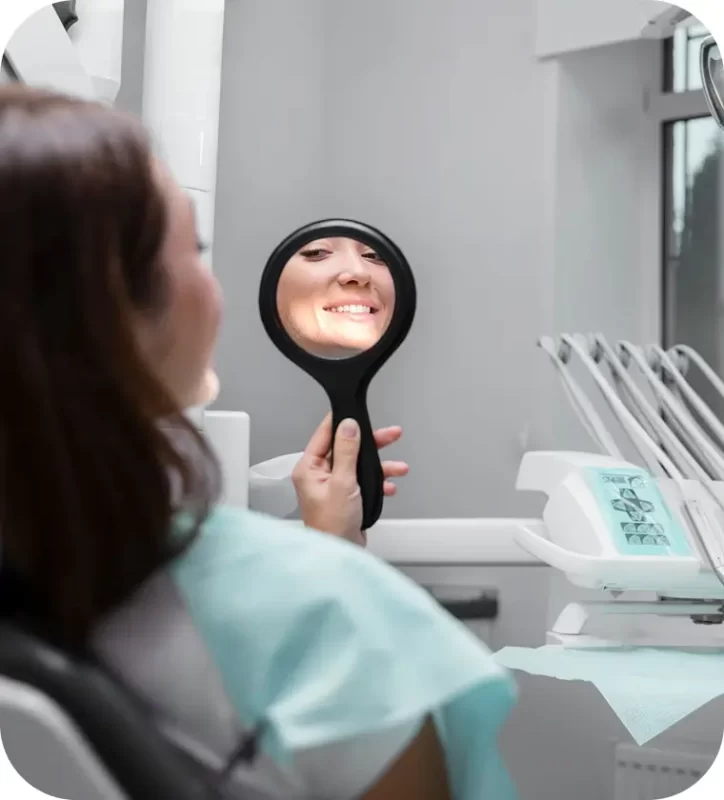 Mujer sonriendo mientras sostiene un espejo en una clínica dental, reflejando resultados de blanqueamiento dental.