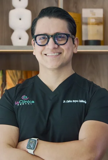 Dr. Carlos Reyes Caldera, dentista de La Clínica Dental, sonriendo con uniforme negro y gafas, fondo moderno y acogedor.