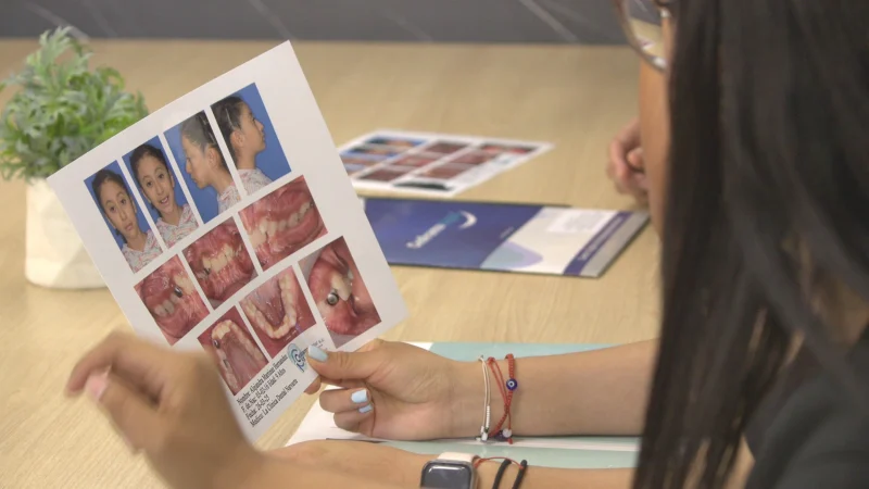 Mujer revisando folleto con imágenes de tratamientos dentales, incluyendo ortodoncia y rehabilitación, en consulta en clínica dental.