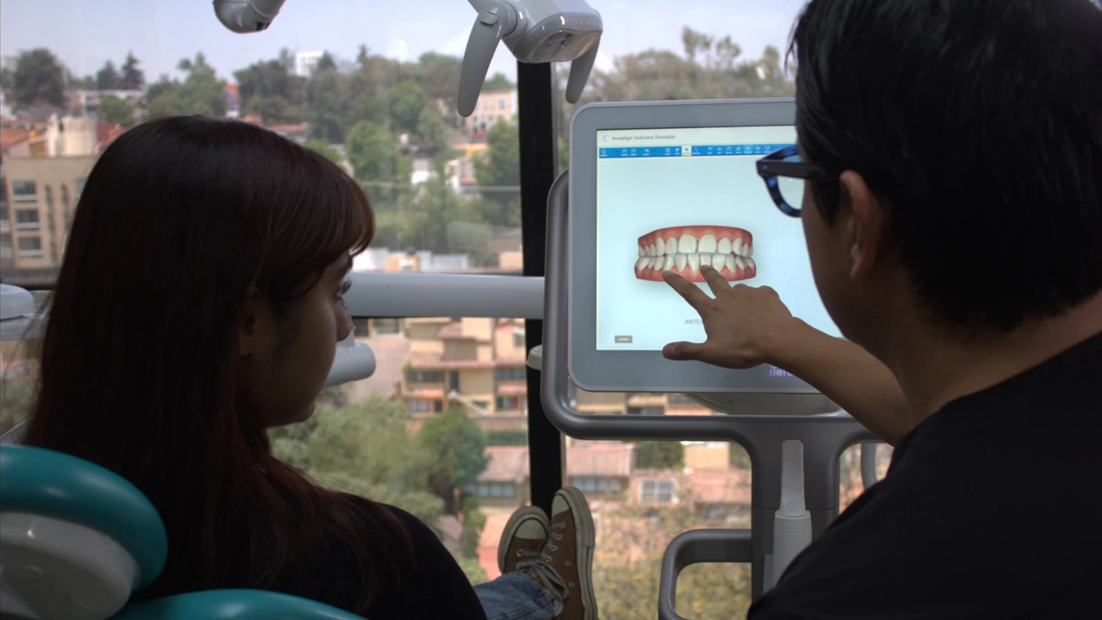 Paciente escuchando a dentista que señala simulador de resultados de tratamiento dental, vista panorámica de CDMX al fondo.
